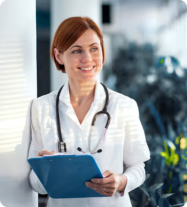 Smiling female doctor in white coat with stethoscope holding a blue clipboard indoors with plants in background.